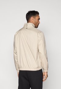 Krēmkrāsas viegla jaka ar augstu apkakli, elastīgiem aprocēm un apakšu, uz muguras neliels logotips un gluda tekstūra.