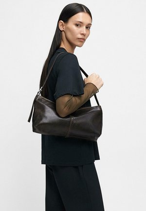 Młoda kobieta o długich ciemnych włosach, ubrana w czarną odzież, trzyma ciemnobrązową skórzaną torbę na ramię na jednolitym tle.