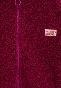 Fleece bunda v tmavo burgundskej farbe s jemným, textúrovaným povrchom; obsahuje fialový zips a nášivku s nápisom "DOBRÉ VECI PRICHÁDAJÚ."
