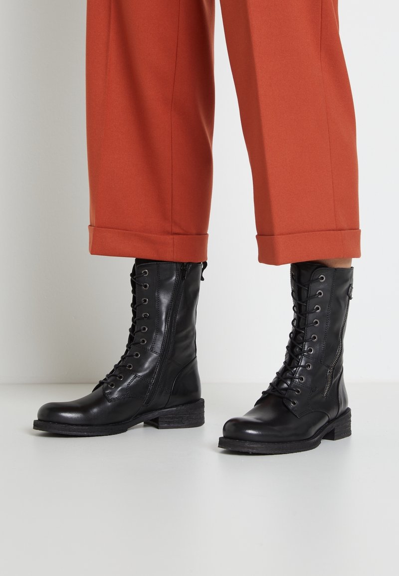 Bottes de combat en cuir noir avec un laçage sur le devant, une finition texturée et une fermeture éclair latérale. Portées avec un pantalon orange retroussé.