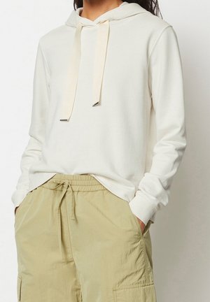 Personne portant un sweat à capuche écru avec des cordons larges et un pantalon cargo beige, les mains dans les poches.