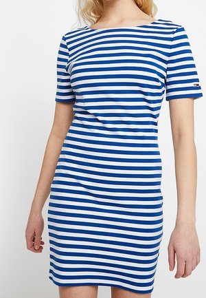 Kvinde iført en kortærmet, blå og hvidt vandret striped kjole med bådhals, stående mod en ensfarvet baggrund.