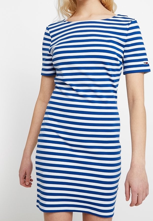 Femme portant une robe à manches courtes à rayures horizontales bleues et blanches avec un col bateau, debout devant un fond uni.