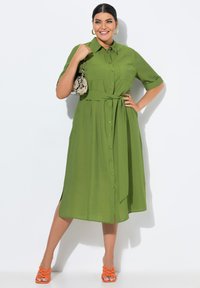 Donna sorridente in abito verde con cintura, che tiene una borsa in pelle di serpente, indossando orecchini a cerchio dorati e sandali arancioni con tacco, in posa.