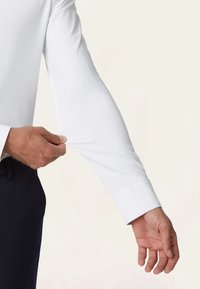 Weißes Langarmhemd mit glatter Textur, taillierter Passform und geknöpften Ärmeln, das an einem Ärmel von einer Hand angepasst wird.