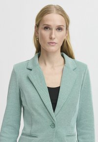 Mintgrön blazer med sned krage, enkel knappstängning och strukturerad tyg. Har framfickor och skräddarsydd passform.