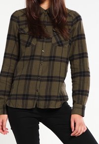 Olivgrön och svart rutig flanellskjorta med knäppning framtill, långa ärmar och två bröstfickor. Mjuk textur, rundad nederkant.