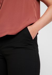 Roze blouse van gladde stof, gecombineerd met zwarte slim-fit broek. Zichtbare zak aan de linkerkant van de broek.