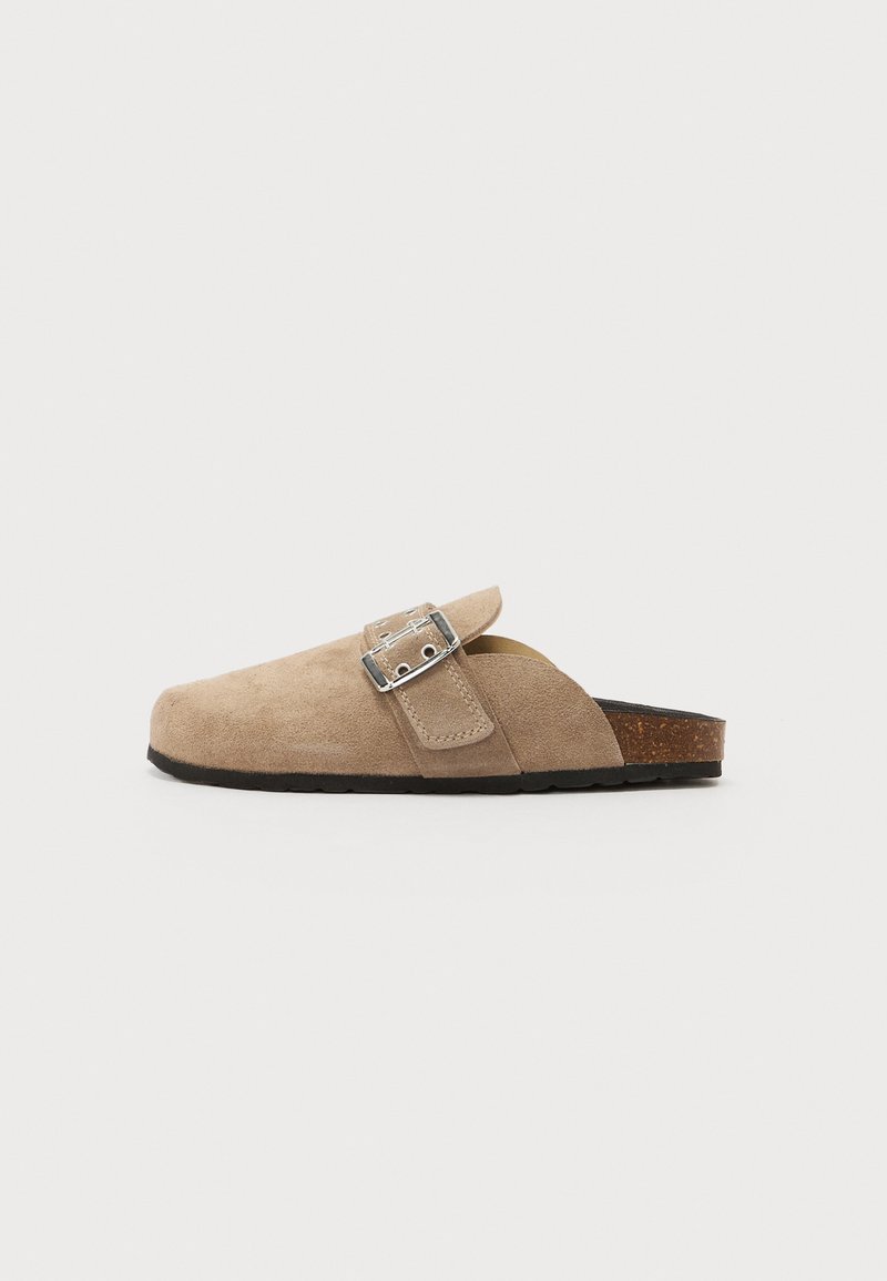 Beige suede slip-on sandal with a buckle strap, rounded toe, and a textured cork footbed. Black rubber outsole. Minimalist design.