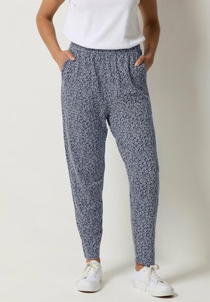 Kvinde iført marineblå bukser med hvidt blomsterprint, hvide sneakers og hvid ærmeløs top, hænderne i lommerne, stående mod ensfarvet baggrund.
