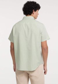 Camicia a maniche corte a quadri verdi con colletto, orlo curvo e tessuto testurizzato, abbinata a pantaloni di colore chiaro.