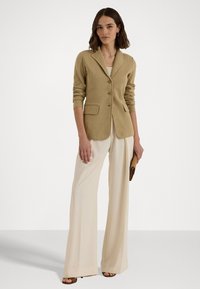 Beige skreddersydd blazer med sjalkrage, tre knapper og to frontlommer, båret over en hvit topp og kombinert med kremfargede vide bukser.