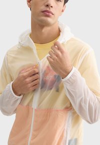Veste coupe-vent légère et claire avec une couche extérieure blanche, des accents pêche et jaune, fermeture zippée et capuche avec cordon de serrage. Détails en mesh présents.