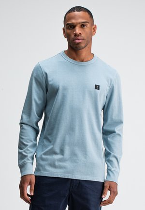 Lichtblauw katoenen overhemd met lange mouwen, met een ronde hals en een klein zwart merklabel op de linkerkant van de borst. Soepele textuur, relaxed fit.