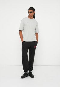 Hombre con cabello trenzado que lleva gafas de sol, camiseta oversized gris claro, pantalones de chándal negros con logo rojo y zapatillas negras, de pie con las manos en los bolsillos.