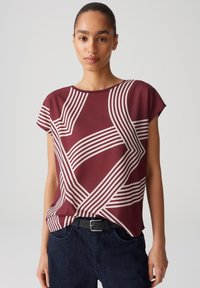 Burgunderfarbenes Kurzarmoberteil mit einem geometrischen weißen Linienmuster. Hergestellt aus glattem Stoff mit Rundhalsausschnitt, getragen über dunklen Jeans.