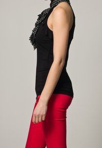 Haut sans manches noir avec un col en volants, associé à un pantalon rouge ajusté. L'écharpe présente un motif à pois, ajoutant un contraste visuel.