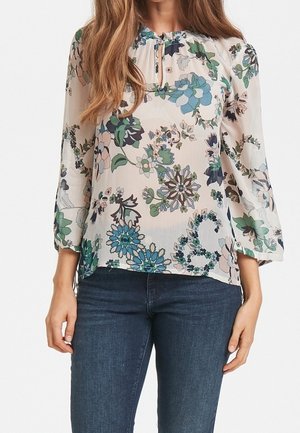 Frau trägt eine transparente, langärmlige Bluse mit floralem Muster in Blau-, Grün- und Beigetönen, kombiniert mit dunkelblauer Jeans.