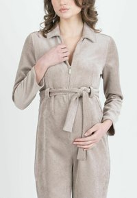 Mono de pana beige con cuello, cierre frontal de cremallera, mangas largas y cinturón anudado en la cintura. Tejido texturizado con patrón de estrías verticales.