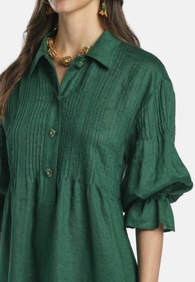 Mujer con blusa verde plisada de mangas abullonadas, botones dorados, un collar grueso de oro y pendientes colgantes coloridos.