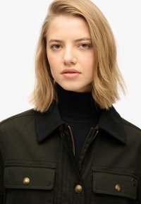 Chaqueta verde oscura con cuello de pana, cremalleras doradas y botones a presión. Cuenta con dos bolsillos en el pecho y una textura acolchada.