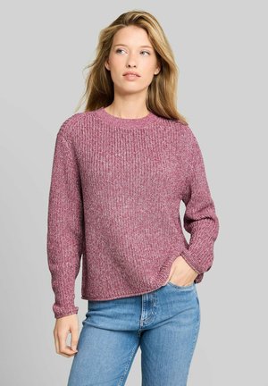 Kobieta z długimi blond włosami, ubrana w różowy sweter z fakturą i niebieskie dżinsy, stoi z jedną ręką w kieszeni na jednolitym tle.