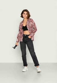 Camisa marmoreada rosa e branca, top preto cropped, jeans pretos de cintura alta e ténis brancos. Modelo segura óculos de sol pretos, em pé contra um fundo claro.