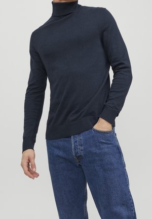 Homme portant un pull à col roulé bleu marine foncé et un jean bleu, une main dans la poche et l'autre détendue le long du corps.