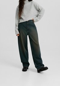 Pull gris oversize à côtes et jean pattes larges vert foncé. Texture tricotée sur le pull ; jean avec un dégradé délavé. Chaussures noires.