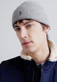 Beanie en tricot côtelé gris avec un petit logo, porté avec une veste bleu marine à col en fourrure. Design épuré et simple, ajusté.