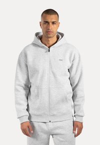 Graue Zip-Hoodie aus weichem Material, mit Kapuze, großen Fronttaschen, gerippten Bündchen und einem kleinen Logo auf der Brust.