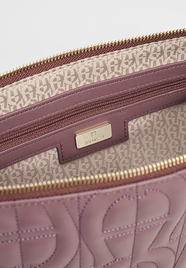 Wnętrze fioletowej skórzanej torby na ramię z gładką teksturą, złotym zamkiem i markową metką. Wyłożone beżową tkaniną z delikatnym wzorem.