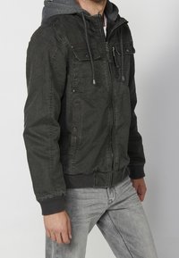 Homme portant une veste zippée vert foncé avec des poches sur la poitrine et une capuche grise, associée à un jean gris clair, debout de profil.