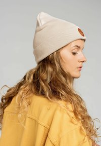 Beżowa dziana czapka z zawiniętym ściągaczem i brązową łatką z logo. Osoba ma długie, faliste włosy i nosi żółtą bluzkę.