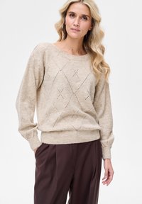 Beżowy sweter na drutach o luźnym kroju, z wzorem diamentowym oraz ściągaczami na rękawach i dole. W zestawie z ciemnobrązowymi spodniami.