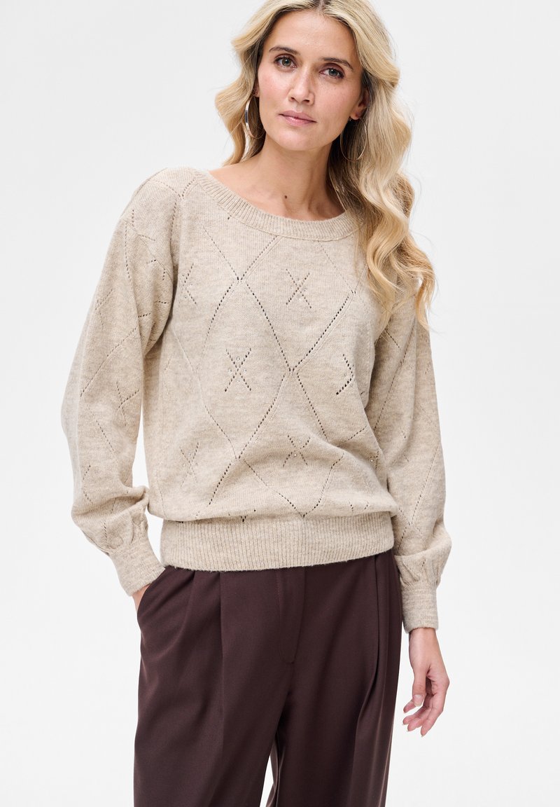 Beżowy sweter na drutach o luźnym kroju, z wzorem diamentowym oraz ściągaczami na rękawach i dole. W zestawie z ciemnobrązowymi spodniami.