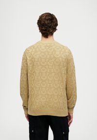 Μπεζ πλεκτό πουλόβερ με ανάγλυφο �σχέδιο λογότυπου, ραμμένες μανσέτες και στρίφωμα, και σχέδιο με στρογγυλή λαιμόκοψη. Μαλακή υφή, χαλαρή εφαρμογή.