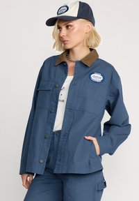 Blaues Arbeitsjackett mit einem Cordkragen, zwei Fronttaschen und Logopatches. Kombiniert mit einer beige-blauen Mütze. Glatte Stoffstruktur.