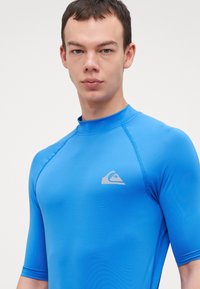 Blå kortermet rash guard laget av glatt, elastisk stoff med rund hals. Har en subtil grå logo på brystet.
