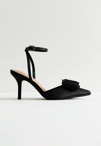 Escarpin noir à bout pointu avec fine bride de cheville et nœud décoratif à l'avant, placé sur un fond blanc uni.