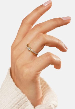 Gold ring with a central gemstone and a band of smaller stones. The hand is shown with a light skin tone and neatly manicured nails.