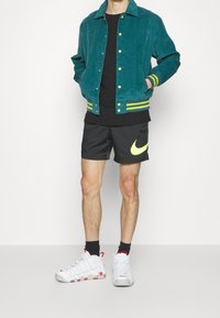 Man med tealjacka med gula accenter, svarta Nike-shorts med neonswoosh, svarta strumpor och vita Nike Air-sneakers med röda detaljer.