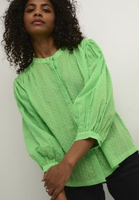 Blouse verte à pois, avec un devant boutonné, un col rond et des manches bouffantes. Tissu transparent et motif texturé avec des détails froncés aux poignets.