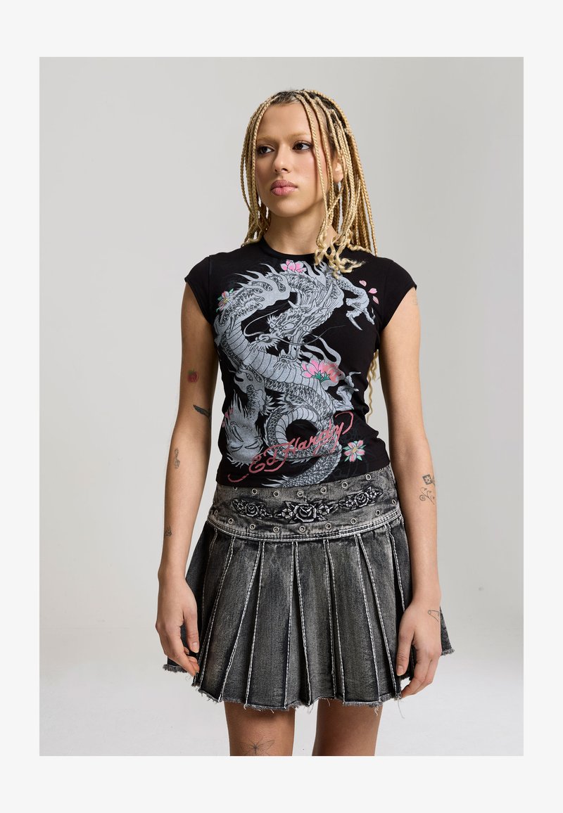 Jongere met lange vlechten, gekleed in een zwart t-shirt met drakenprint en een zwarte geplooide spijkerrok met rozenborduursel.