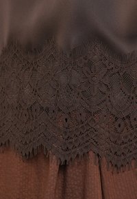 Καφέ ύφασμα με περίτεχνη δαντέλα στο τελείωμα, με σχέδια λουλουδιών και κρόσσια στην άκρη. Η βάση έχει υφή με ρίγες.