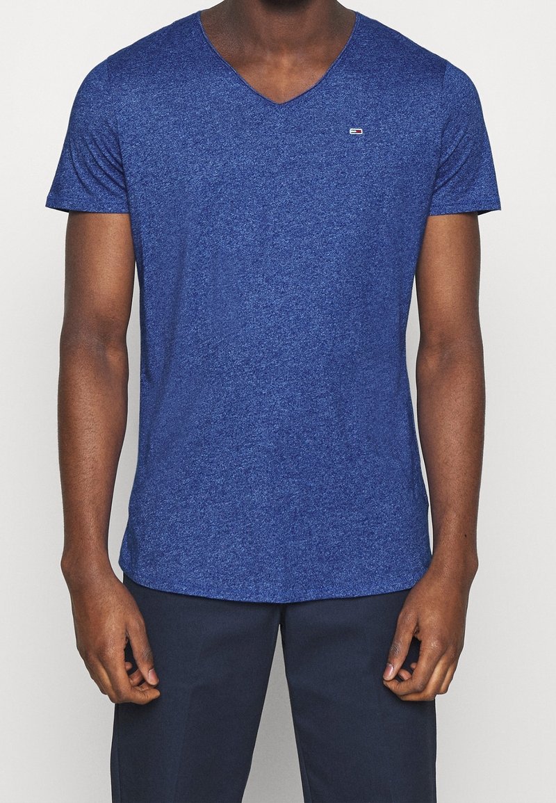 Homme portant un T-shirt V en bleu chiné avec un petit logo sur la poitrine et un pantalon navy foncé, se tenant les bras détendus.