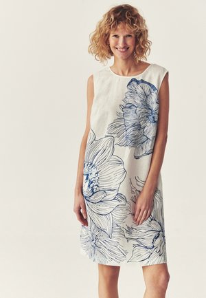 Uśmiechnięta kobieta o kręconych blond włosach, ubrana w białą sukienkę bez rękawów z dużymi niebieskimi kwiatowymi wzorami, stoi na tle jednolitego tła.