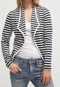 Blazer a righe blu navy e bianco, con un design frontale curvato, colletto aperto e rifiniture a contrasto. Indossato sopra una maglietta bianca e jeans di denim.