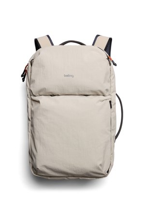 Sac à dos beige clair avec une texture en tissu doux, des fermetures éclair et une poignée supérieure. Dispose de bretelles rembourrées et d'un logo de marque discret à l'avant.