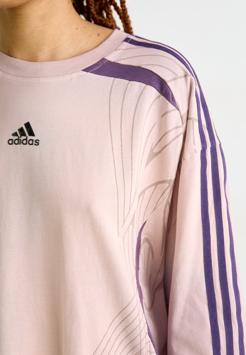 Jasnoróżowa koszula z długim rękawem o subtelny wzorze fal, z ciemnofioletowymi paskami na rękawach i czarnym logo Adidas na piersi.